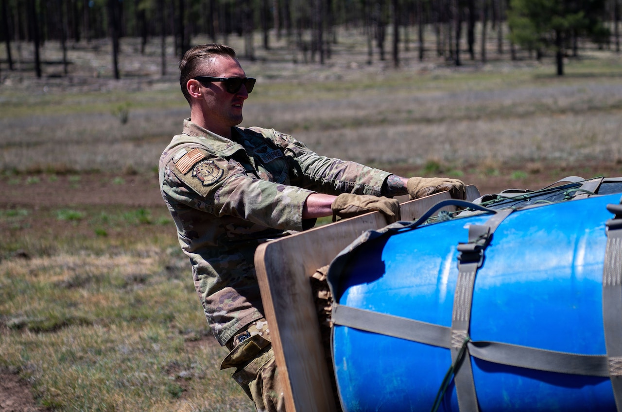A man in a camouflage military uniform and sunglasses moves cargo in an open field that was dropped by a military aircraft.