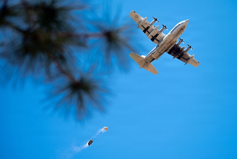 A large military aircraft flies overhead in blue skies as it drops cargo that is parachuting to the ground; branches of a pine tree are blurred in the foreground.