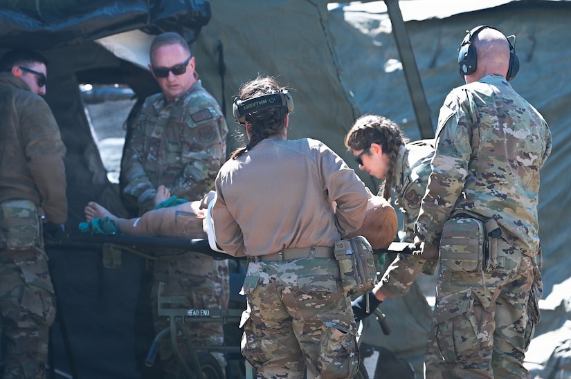 Five people in camouflage military uniforms move a mannequin on a stretcher into a medical tent.