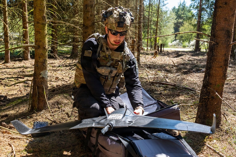 A man wearing a camouflaged military vest and helmet crouches down in the woods while holding a drone.