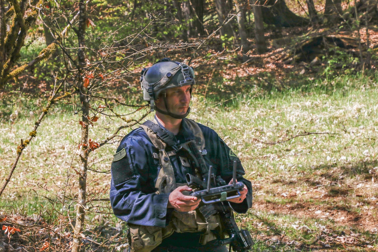 A man wearing a dark blue military uniform, helmet and camouflage vest holds a remote control in the woods and looks at something out of frame.