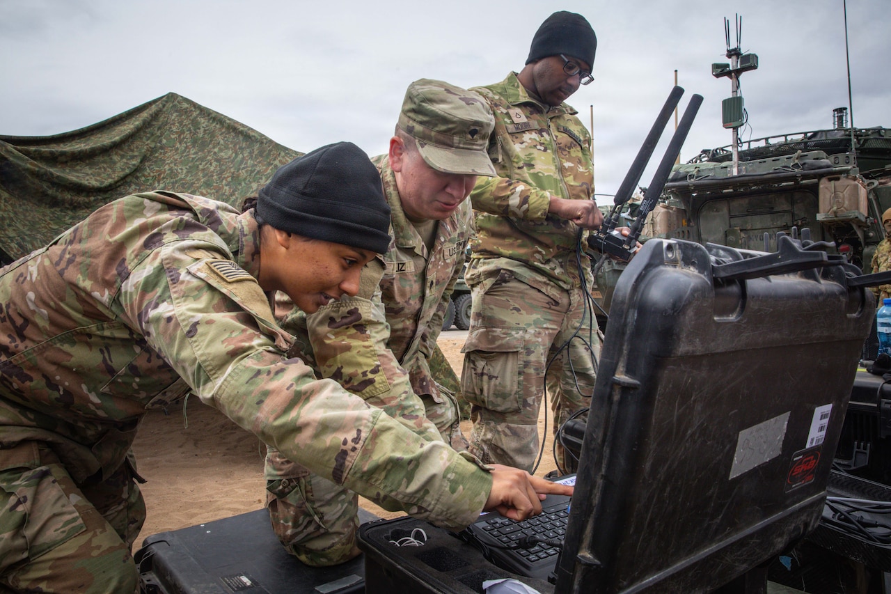 A woman in a camouflage military uniform points to a computer screen sitting on top of a case as a man in similar attire looks on; another man in similar attire is standing in the background holding a piece of military equipment.