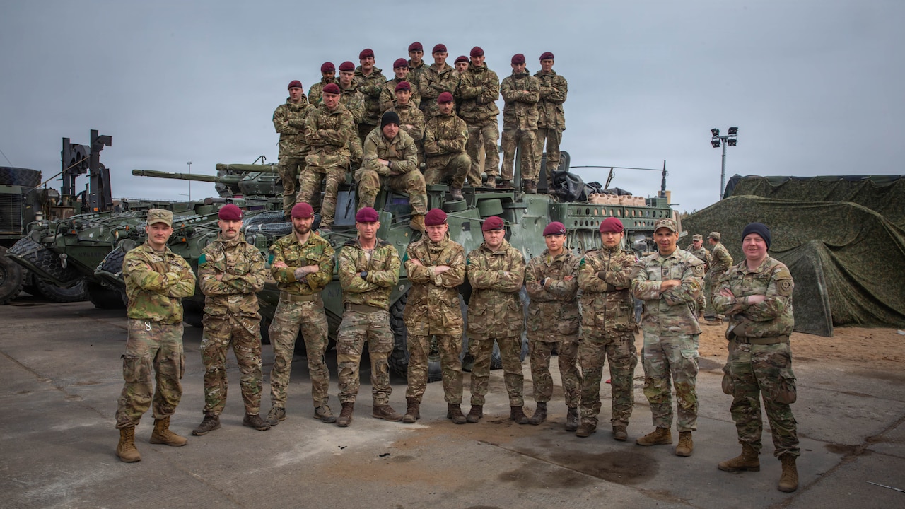 Ten men in camouflage military uniforms pose for a photo in front of a military vehicle while a dozen other men in similar attire stand/sit on top of the vehicle.
