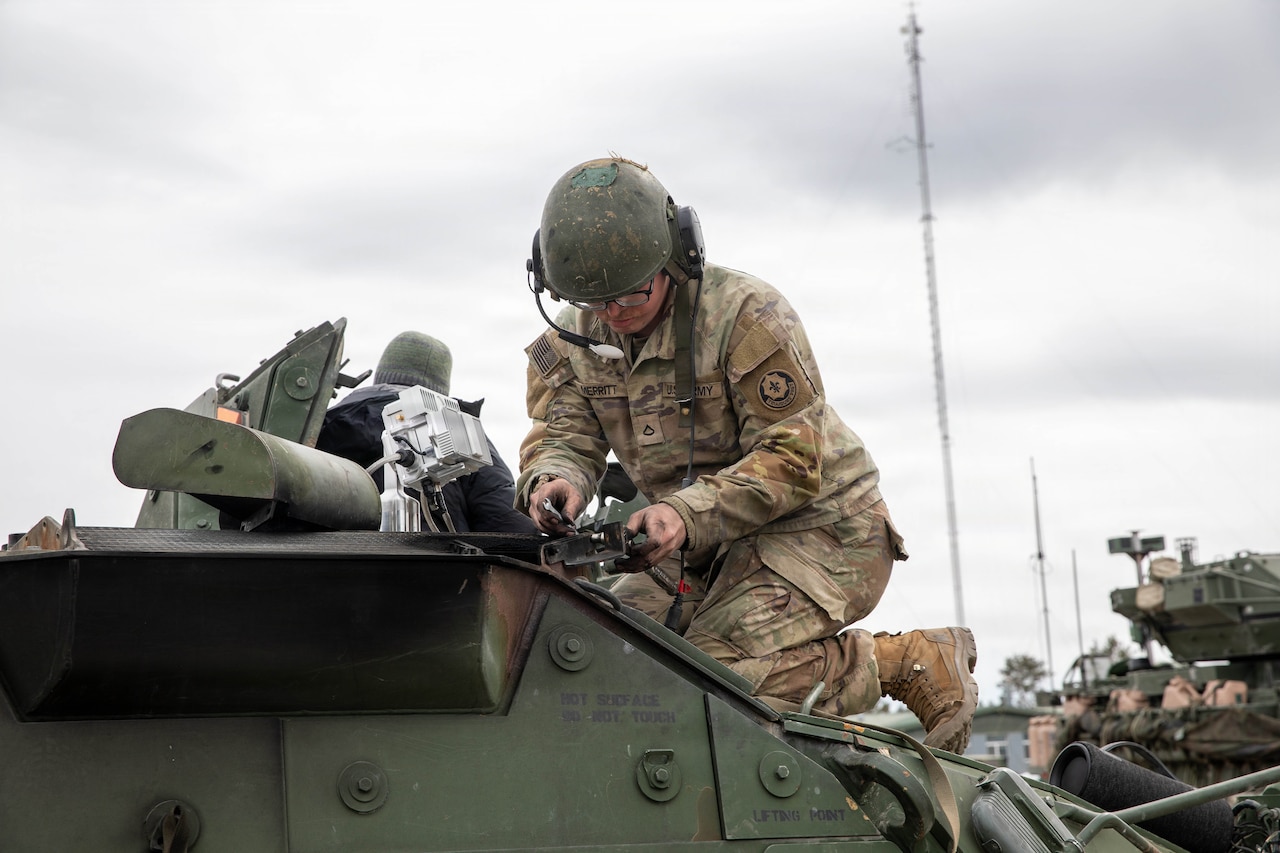 A man in a camouflage military uniform and helmet fixes equipment while kneeling on top of a military vehicle.