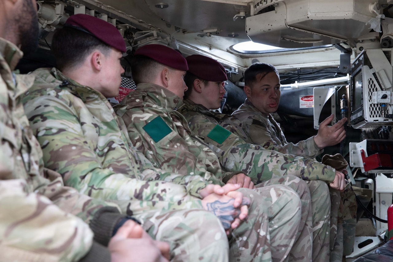 A man in a camouflage military uniform shows how to use a computer system on a military vehicle to four other men in similar attire sitting in the vehicle with him.