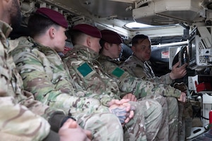 A man in a camouflage military uniform shows how to use a computer system on a military vehicle to four other men in similar attire sitting in the vehicle with him.
