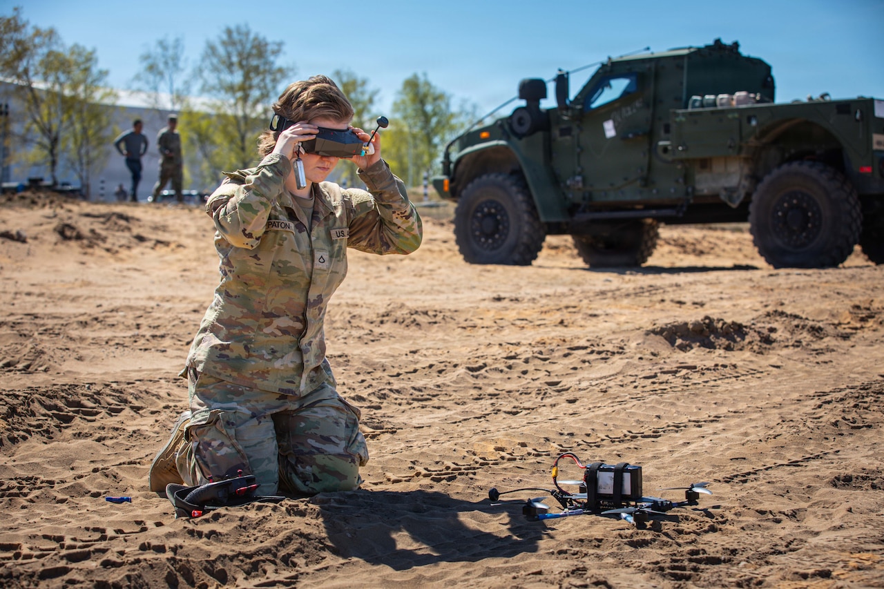 A woman in a camouflage military uniform kneels in the dirt as she places goggles over her eyes with a remote and a drone sitting in the dirt next to her; there is a military vehicle and two other people in similar attire in the background.