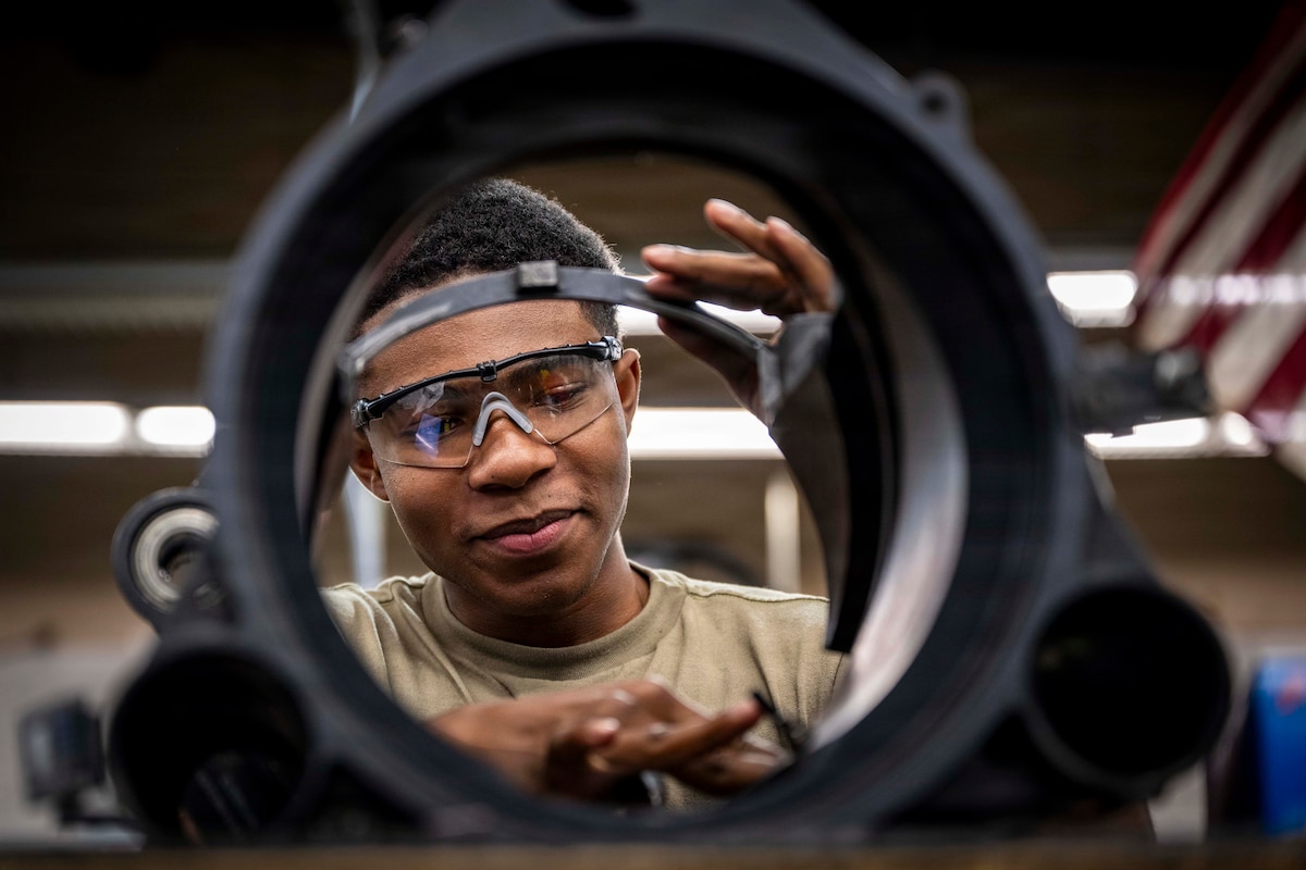 An airman wearing protective glasses works on a circular metal component in a workshop as lights shine in the background.