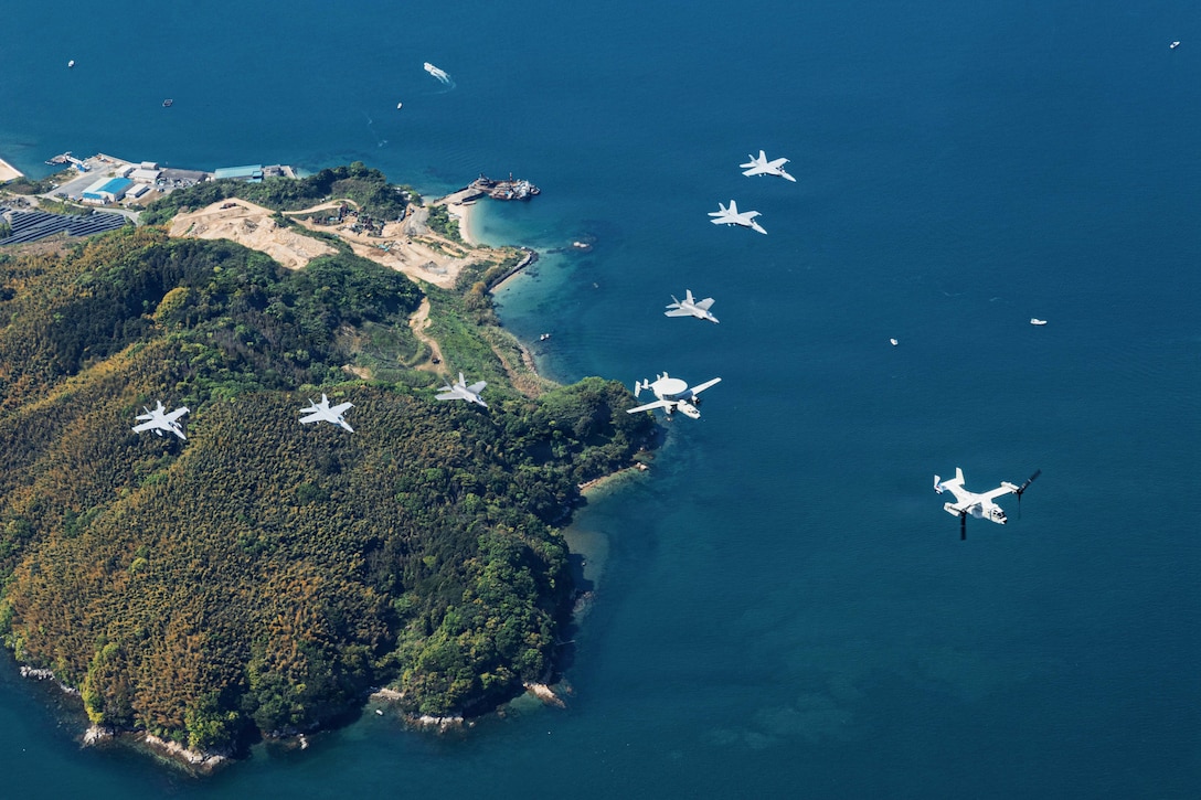 An aircraft leads seven other aircraft flying in a triangular formation over blue waters near an island during the day.