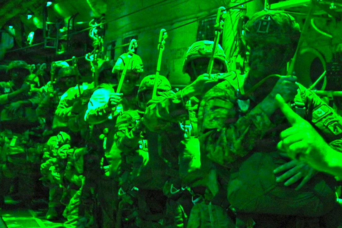 Airmen in tactical gear line up in an aircraft as seen through a night vision lens.