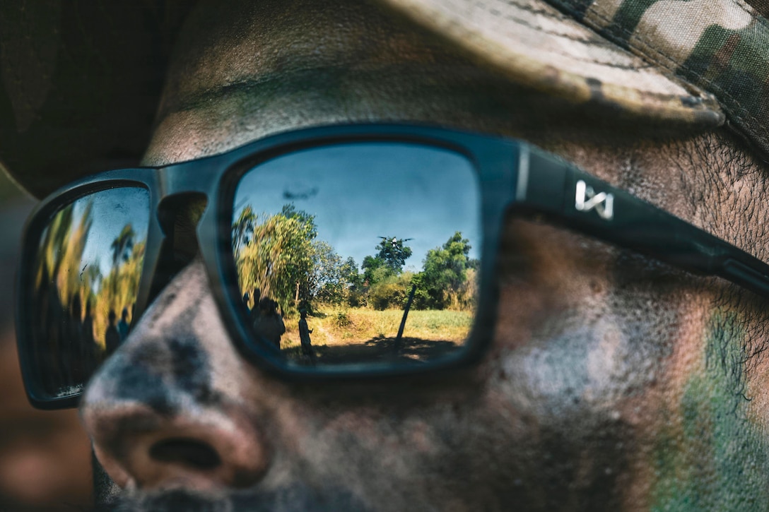 A drone flies near trees in a field as seen through the lens of a soldier in camouflage.