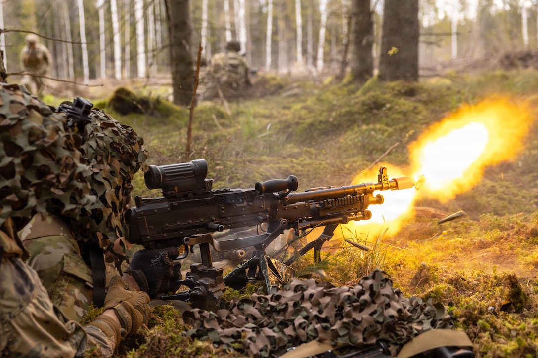 A soldier in camouflage fires a weapon in a field, creating an orange fireball.