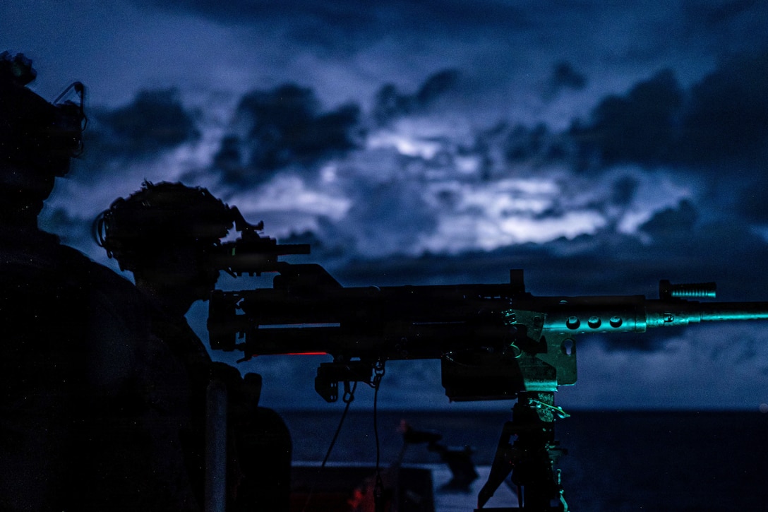 A sailor looks through scope of a weapon in the dark illuminated by a red light.