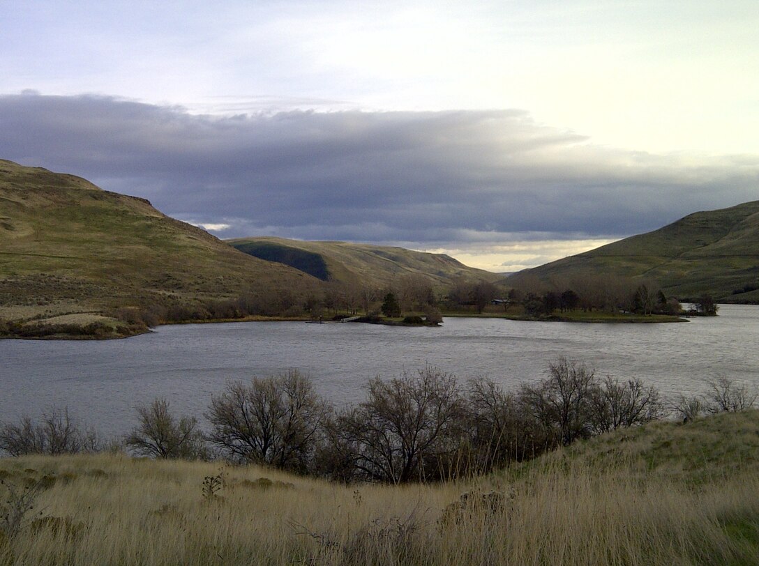 A calm lake nestled between rolling, grassy hills under a cloudy sky, with leafless trees along the shoreline and a small island or peninsula extending into the water.