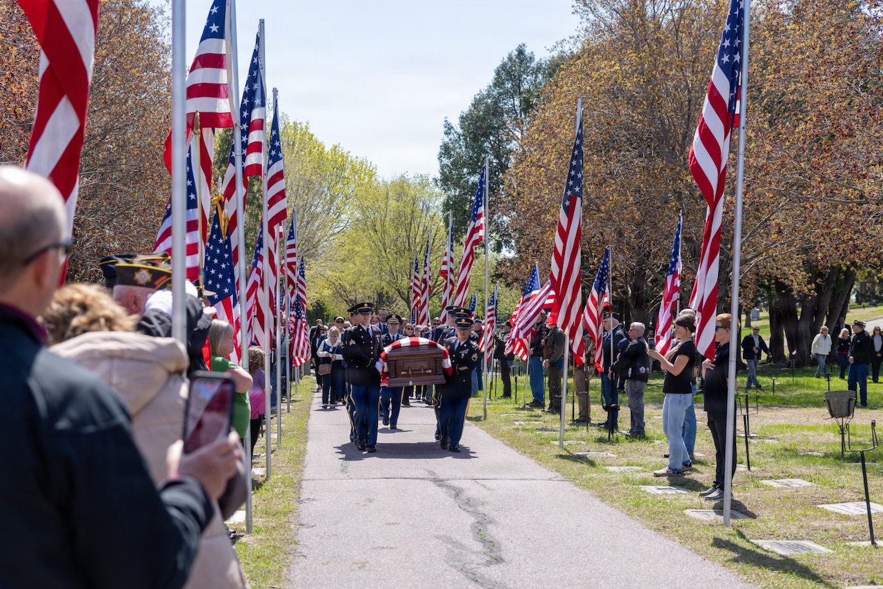People in formal military uniforms carry a casket draped in an American flag down a path lined with people holding American flags in a cemetery.