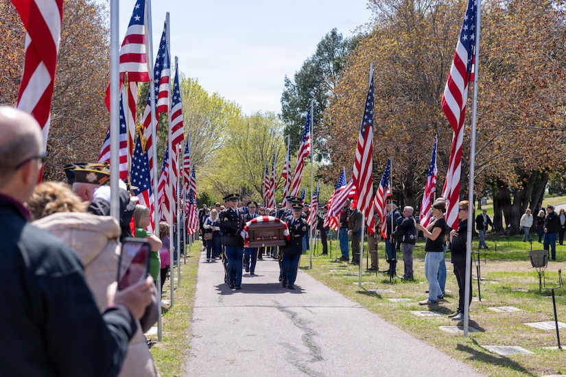 People in formal military uniforms carry a casket draped in an American flag down a path lined with people holding American flags in a cemetery.