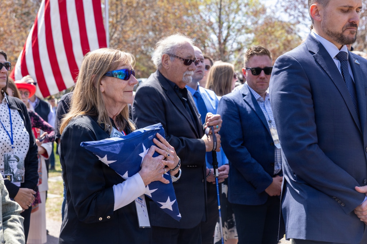 A woman in business attire and sunglasses holds a folded American flag during a ceremony outside, surrounded by people in similar attire.