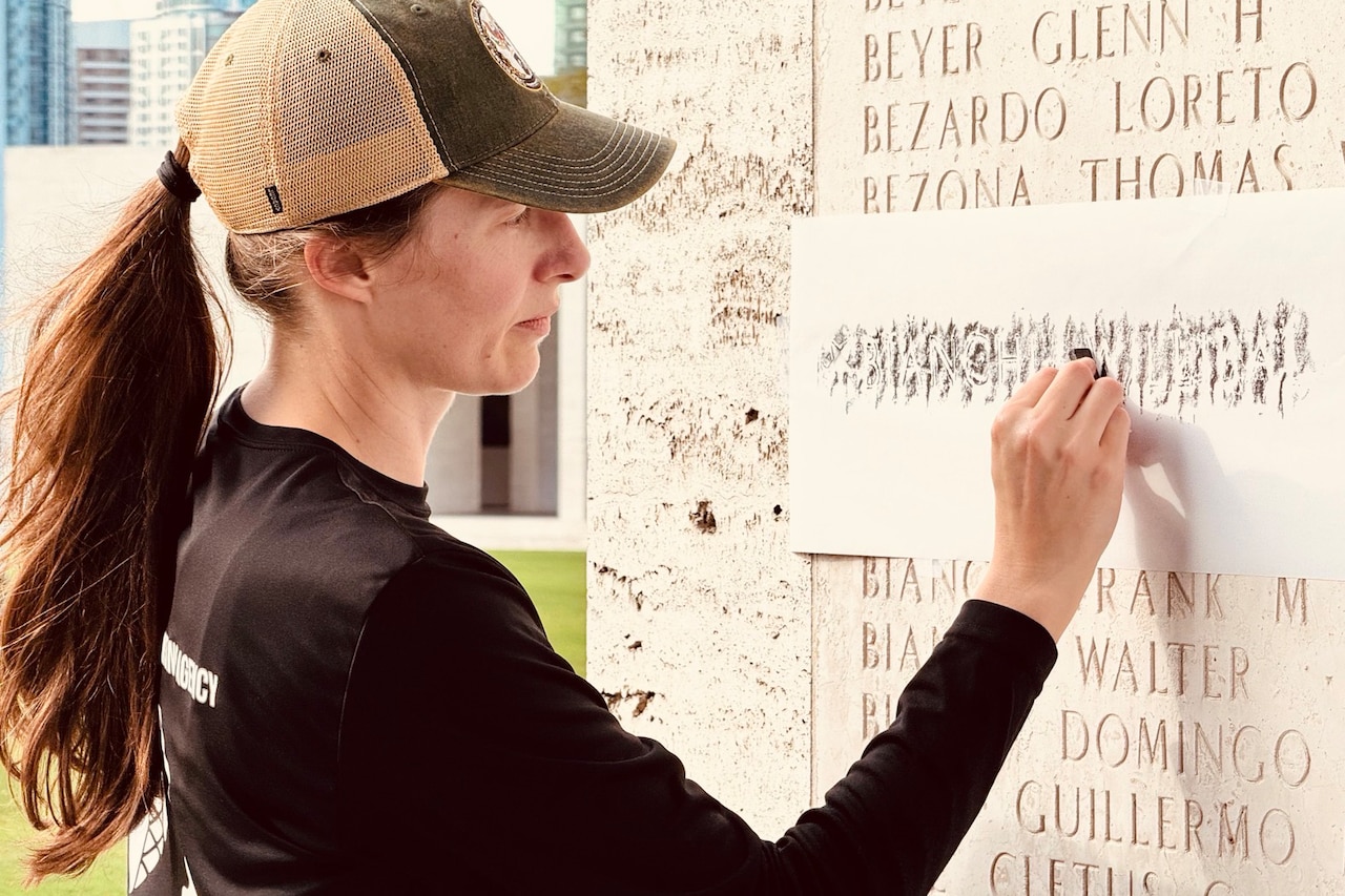 A woman in casual attire stands in front of a stone wall with names engraved and transfers a name from the wall to a sheet of paper.