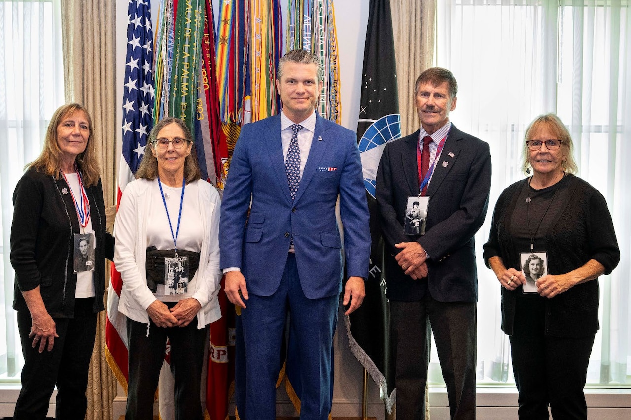 Four people in business attire pose for a photo in front of windows and flags with streamers attached.