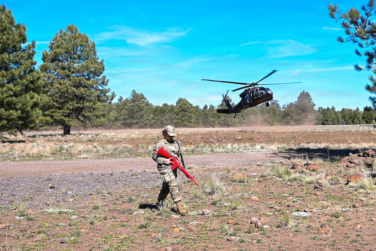 A woman in a camouflage military uniform walks in an open field carrying a training rifle as a military helicopter lands in the background.