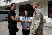U.S. Space Force Brig. Gen. Brian Denaro, in uniform, shakes hands with a man in a black shirt while a woman stands smiling between them. The group is standing outdoors in front of a building.