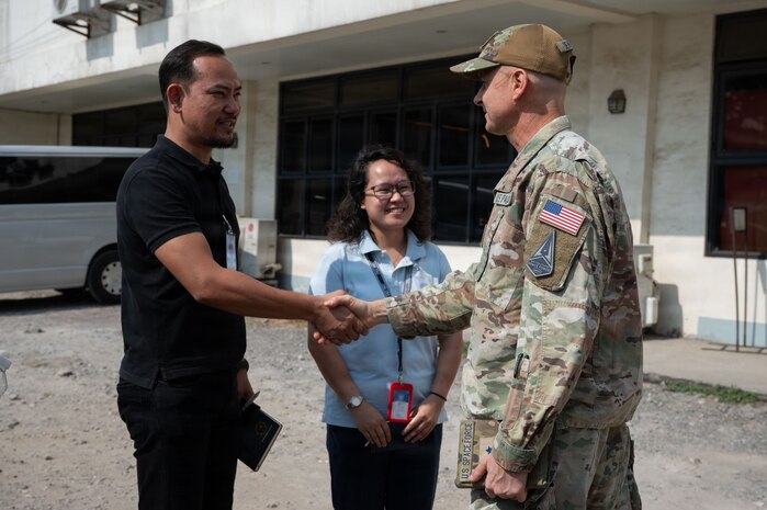 U.S. Space Force Brig. Gen. Brian Denaro, in uniform, shakes hands with a man in a black shirt while a woman stands smiling between them. The group is standing outdoors in front of a building.