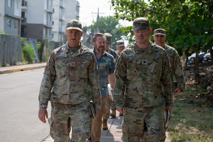 U.S. Space Force Brig. Gen. Brian Denaro and Lt. Col. Lee Schroeder, both in uniform, walk down a street leading a group of people. A man in a patterned, short-sleeved shirt is visible walking just behind them with the rest of the people following in military uniforms, with residential buildings in the background.