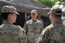 U.S. Space Force Brig. Gen. Brian Denaro, in uniform, smiles while speaking with two other service members in front of a tent with a textured net on top.