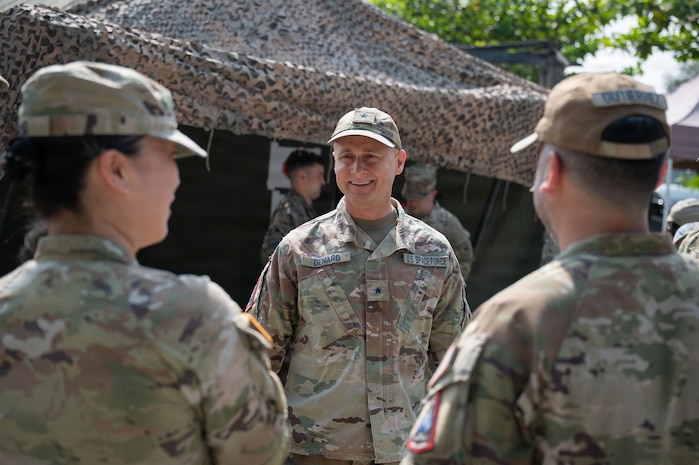 U.S. Space Force Brig. Gen. Brian Denaro, in uniform, smiles while speaking with two other service members in front of a tent with a textured net on top.