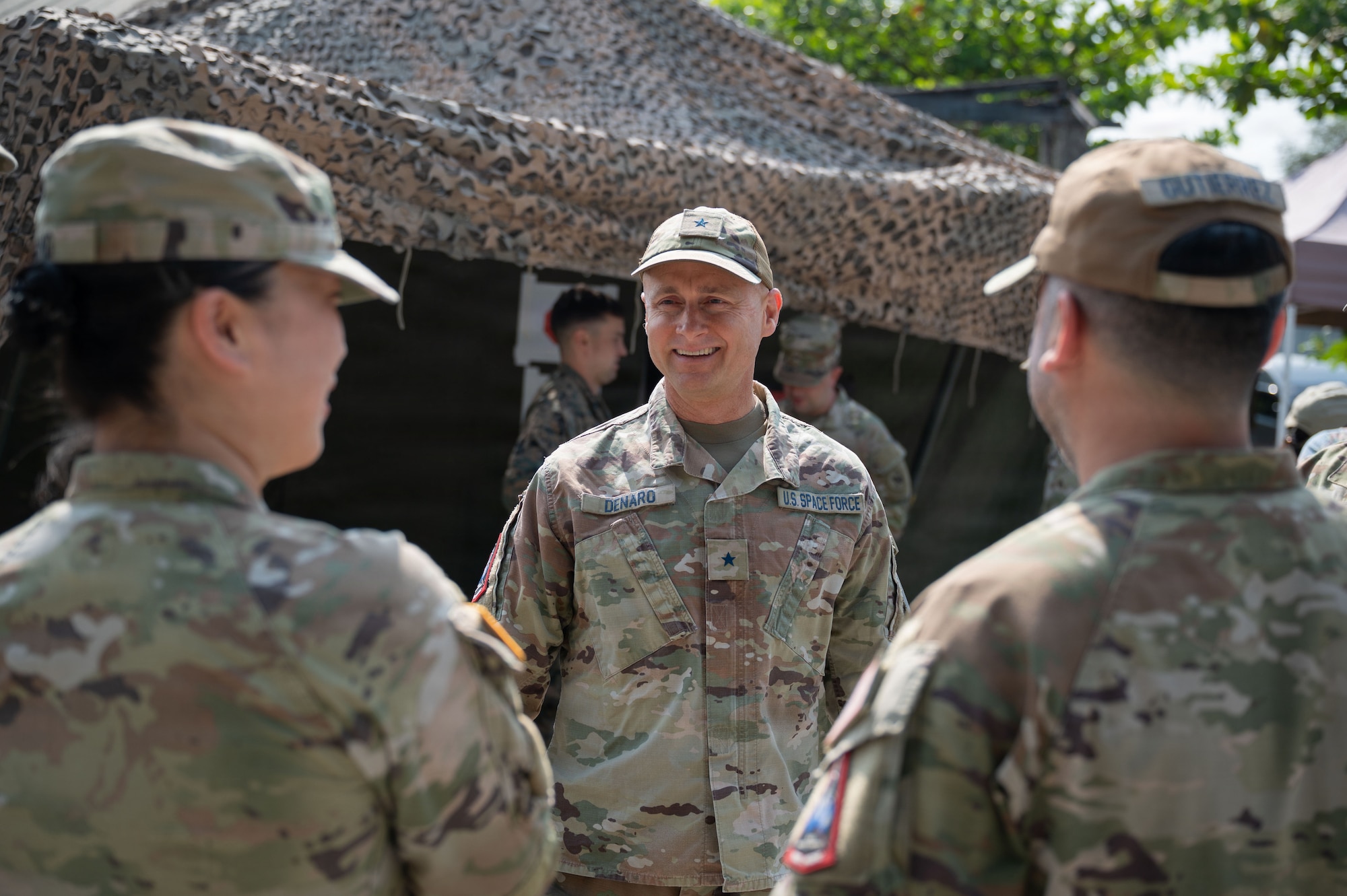 U.S. Space Force Brig. Gen. Brian Denaro, in uniform, smiles while speaking with two other service members in front of a tent with a textured net on top.