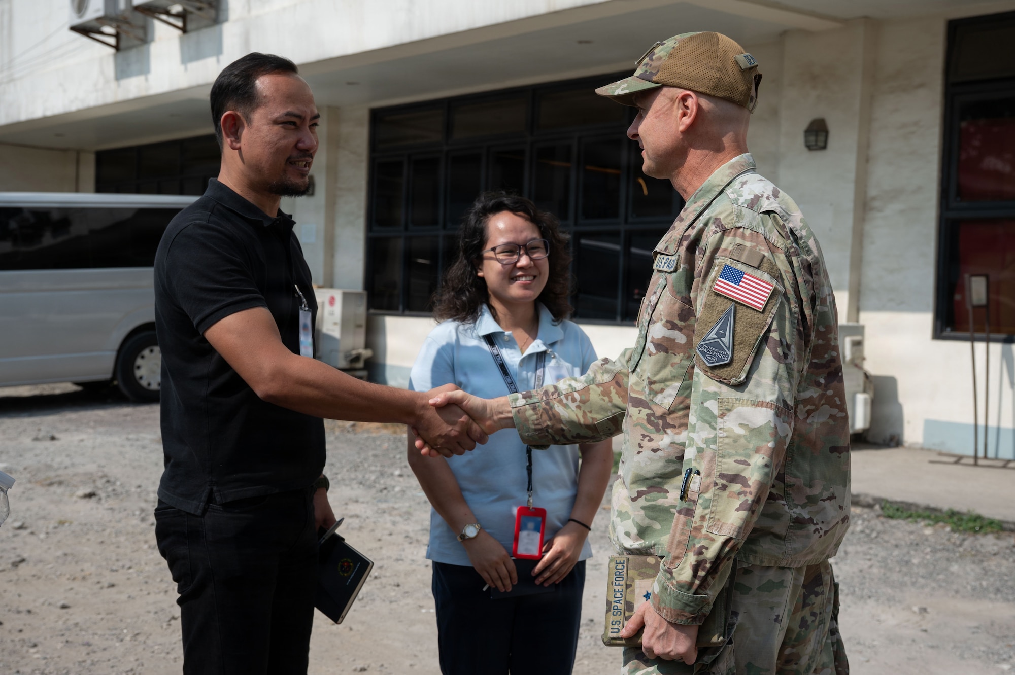 U.S. Space Force Brig. Gen. Brian Denaro, in uniform, shakes hands with a man in a black shirt while a woman stands smiling between them. The group is standing outdoors in front of a building.