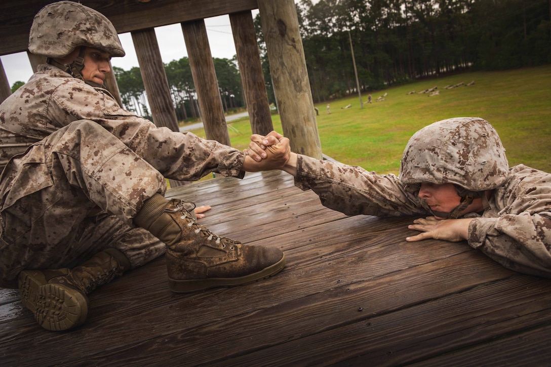A seated Marine Corps recruit pulls a fellow recruit up a wooden obstacle with a wooded area in the background.