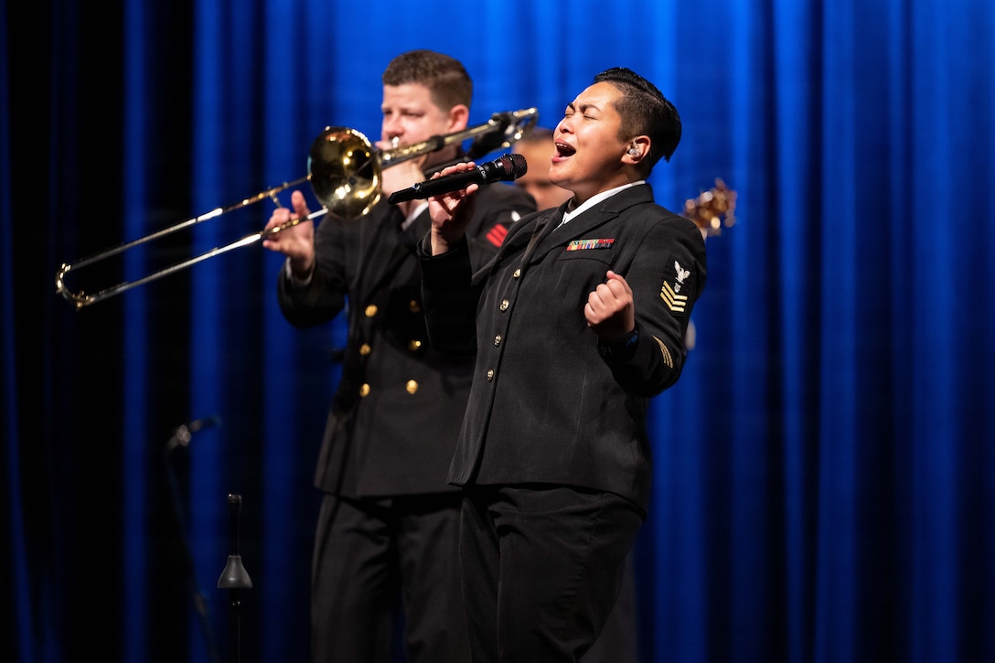 A sailor uses a microphone to sing near fellow sailors playing instruments against blue curtains.