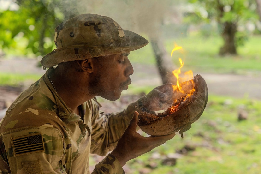 A soldier blows on a burning coconut against a wooded area.