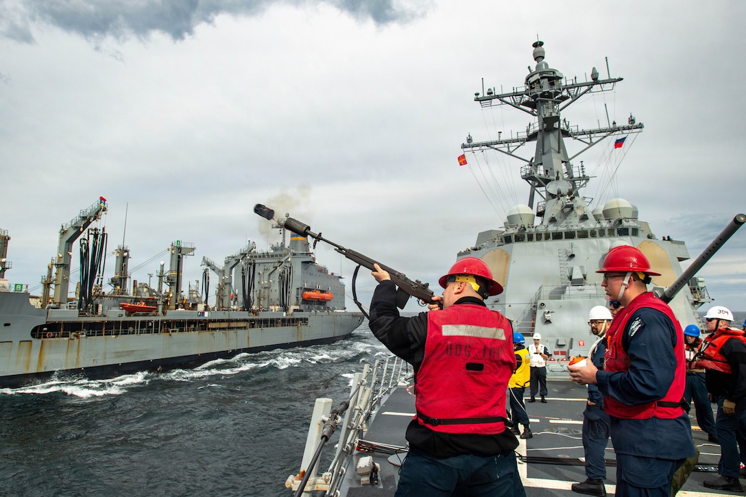 Sailors watch as a sailor in a red vest fires a shot line from a ship to another ship on a gloomy day.