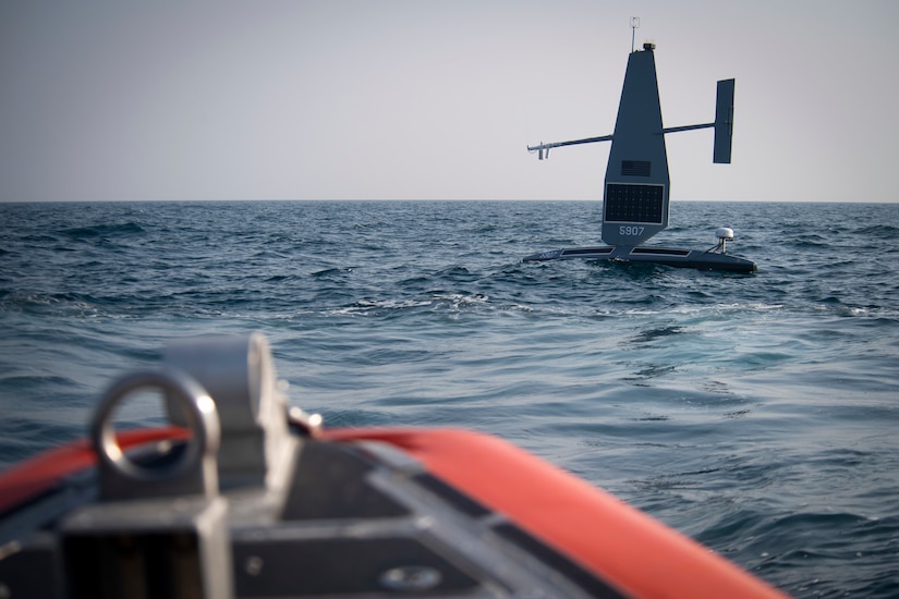 An unmanned military vessel is shown floating in a body of water. The bow of another vessel is shown in the foreground.