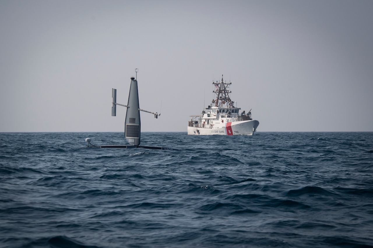 An unmanned military vessel is shown floating in a body of water. A military ship is in the background.