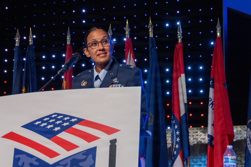 A woman in a military dress uniform speaks on a stage standing behind a lectern; there are flags displayed in the background.