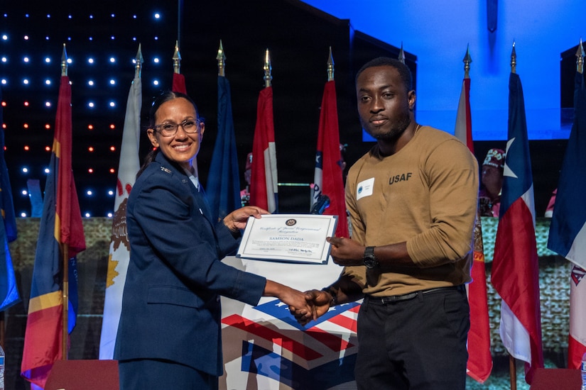 A woman in a military dress uniform and a man in casual attire pose for a photo while holding a certificate in their hands; there are flags displayed in the background.