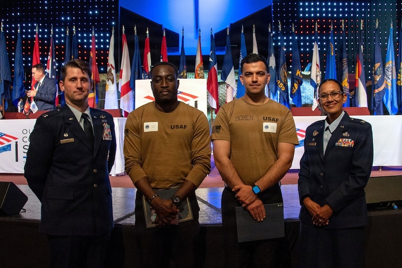 A man and a woman in military dress uniforms pose for a photo in front of a stage with two men in casual attire holding certificates; there are flags displayed in the background.
