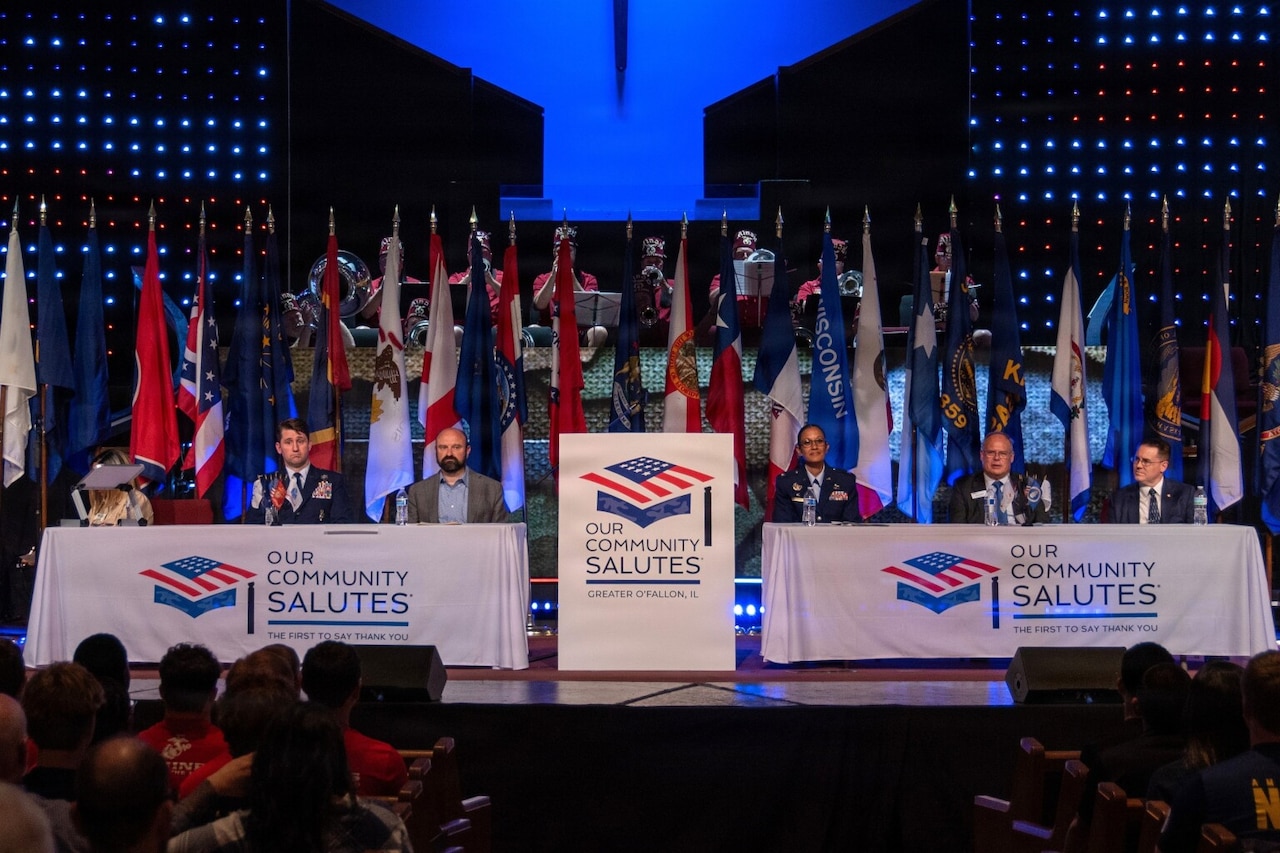 Six people in business attire and military dress uniforms sit behind two tables on a stage during a military event; there are flags displayed in the background. A lectern in the center of the stage and the two tables featured logos that read, “Our Community Salutes.”