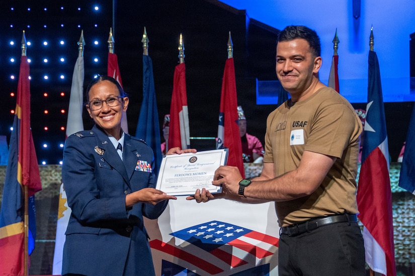 A woman in a military dress uniform and a man in casual attire pose for a photo while holding a certificate in their hands; there are flags displayed in the background.