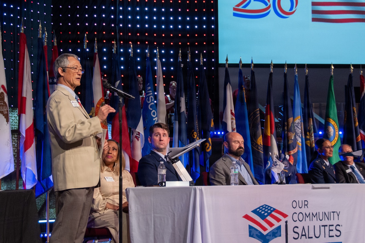 A man in a suit speaks into a microphone while standing on a stage during a military event; there are five people in business attire and military dress uniforms seated on stage to the right with flags in the background.