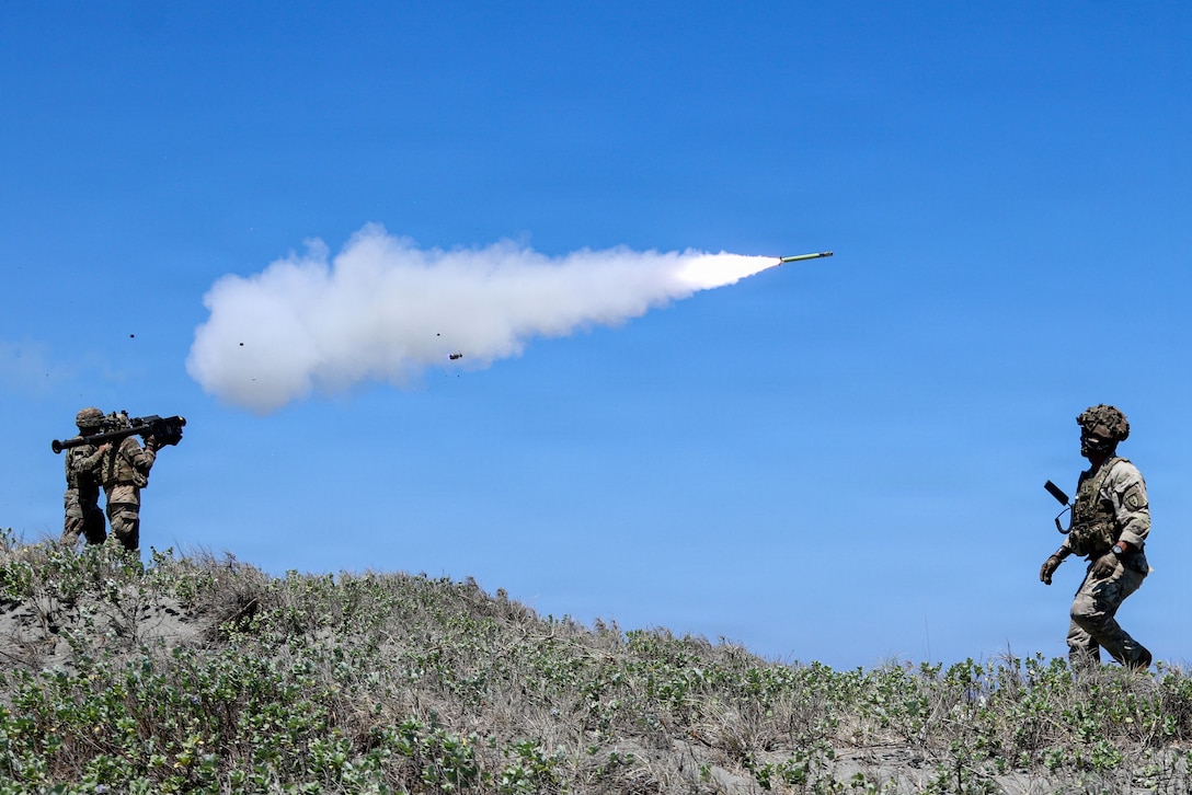 Two soldiers hold a device and fire a rocket on a hillside, as another soldier walks in the distance and looks at the rocket and smoke in the air.