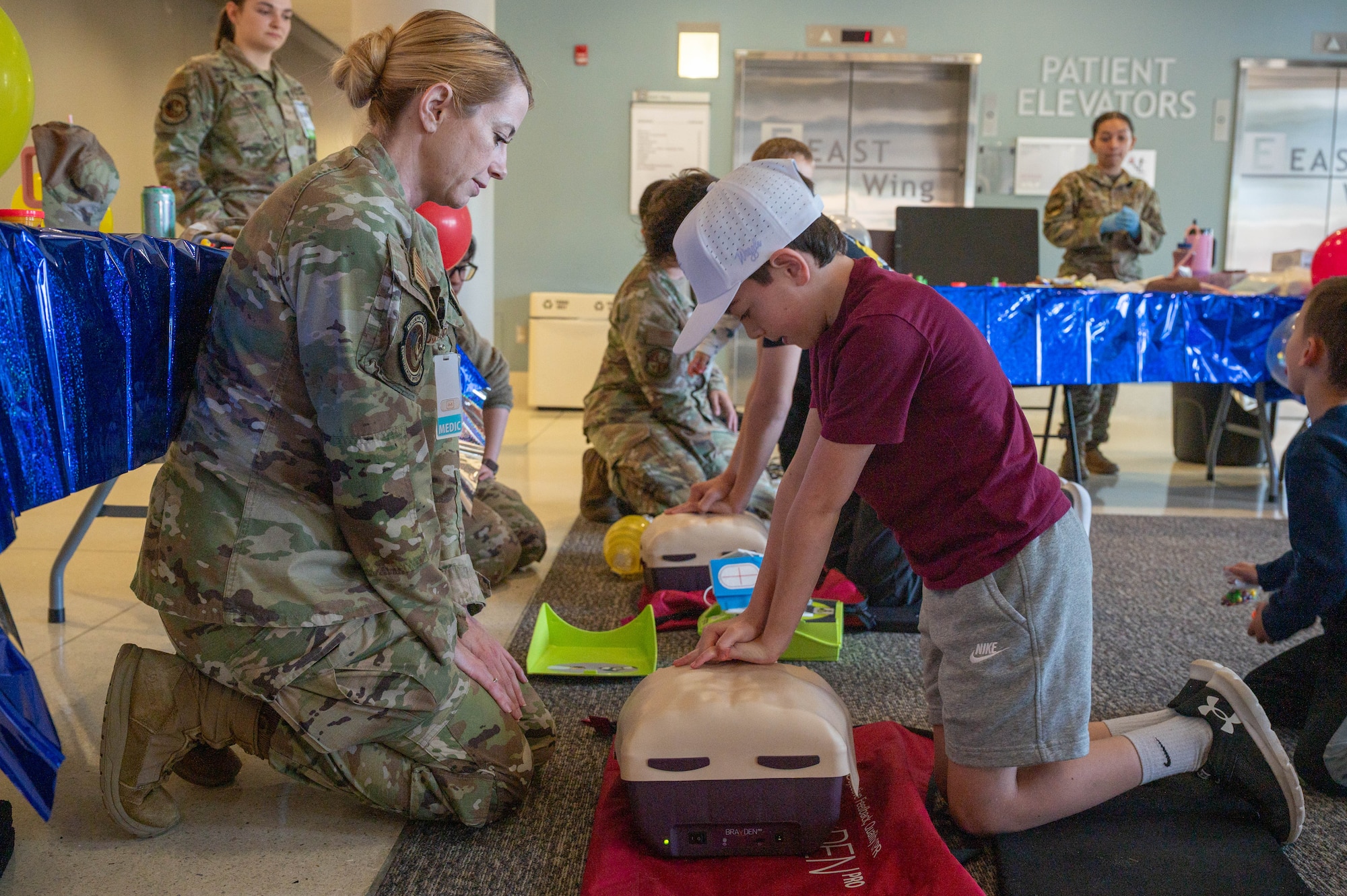 Photo shows a child on the right performing simulated compressions on a mannequin with an Airman on the left watching.