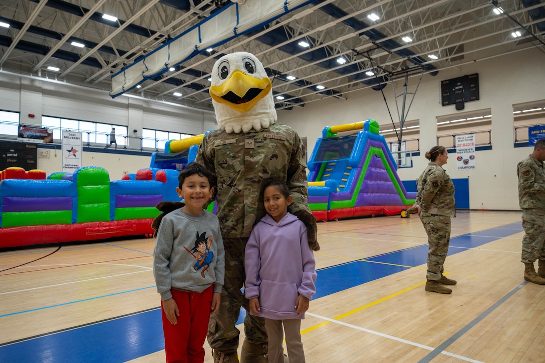Photo shows JB, the JBLE eagle mascot, standing with two children, posing for a photo with bounce castles in the background.