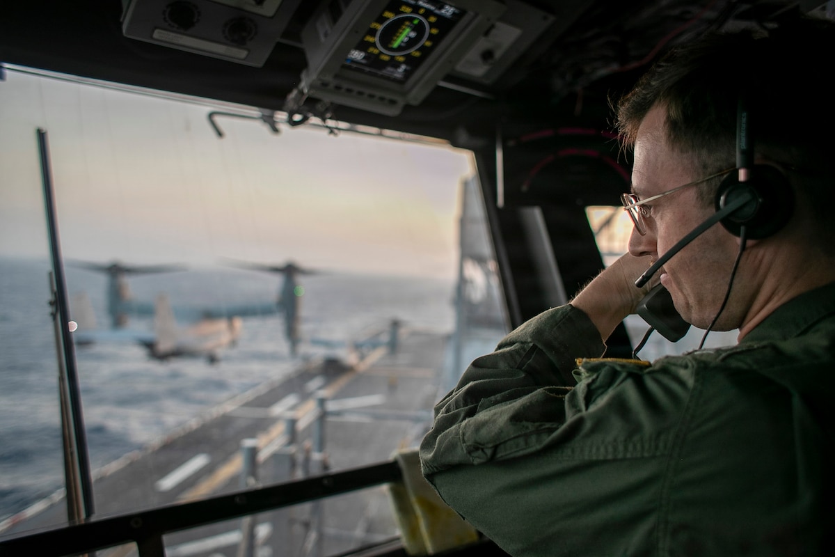 A sailor looks out of the window of a control tower on a ship at sea, with an aircraft hovering in the background during daytime.