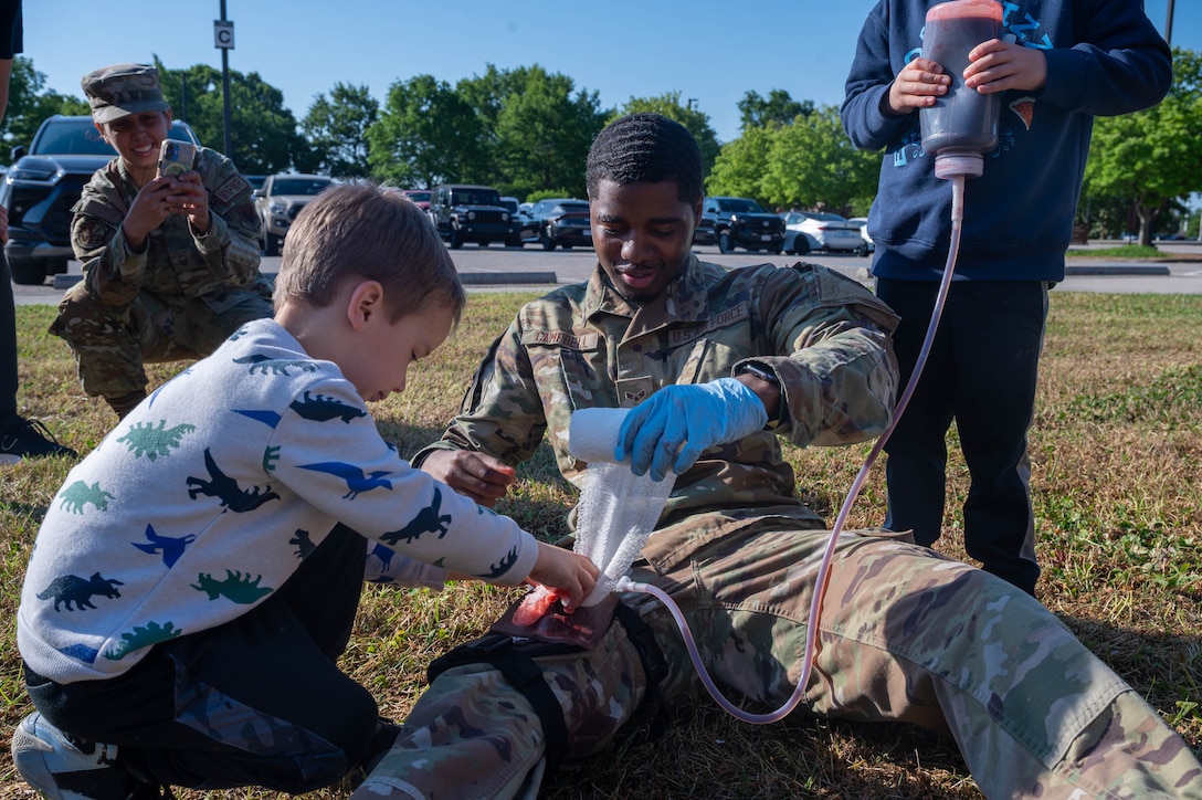 Photo shows a child on the left packing a simulated wound on an Airman's leg.