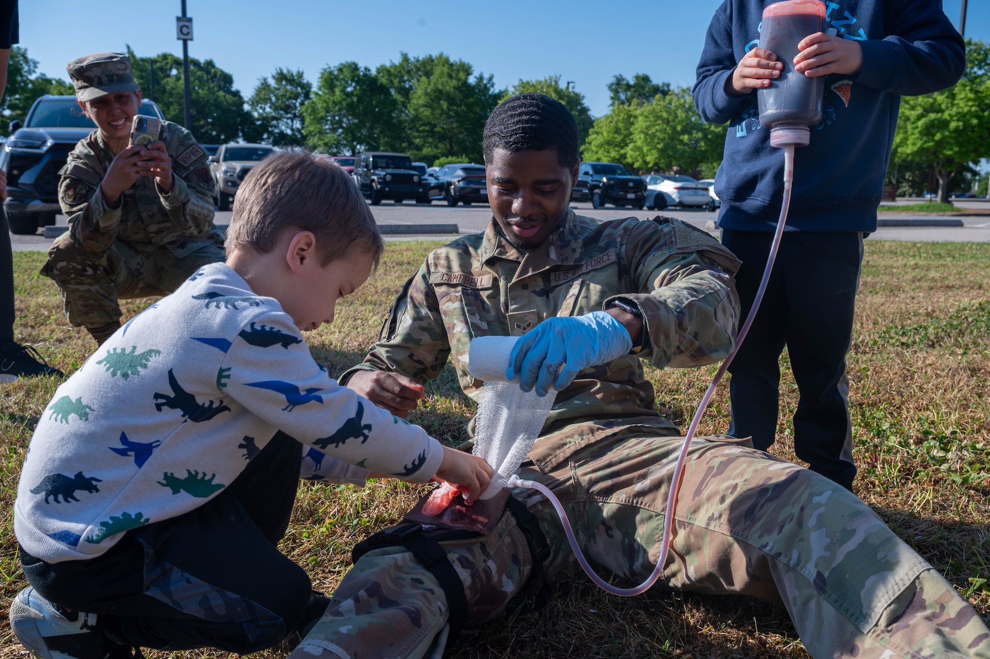 Photo shows a child on the left packing a simulated wound on an Airman's leg.