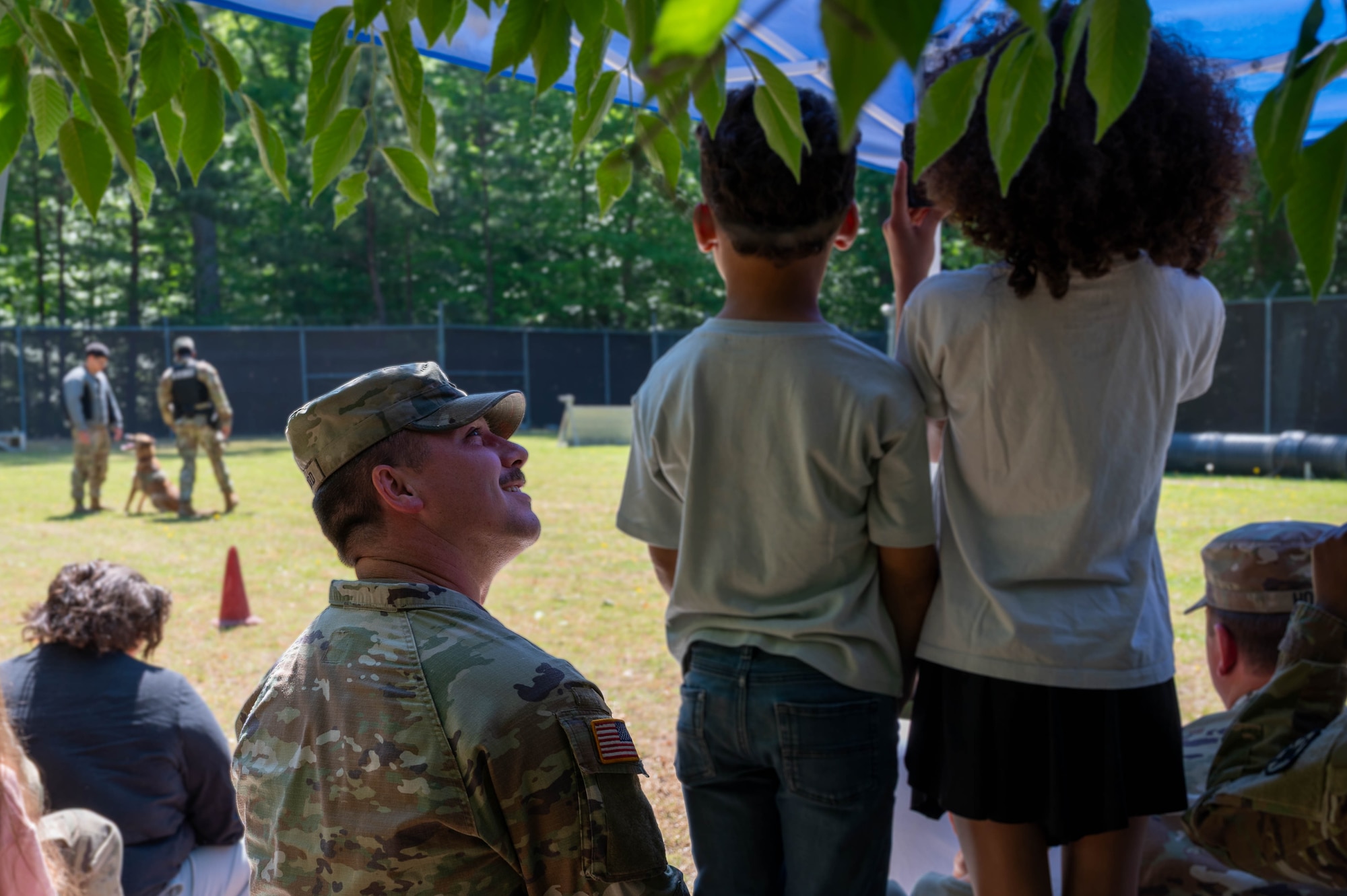 Photo shows a military working dog demonstration in the background as a military dad smiles as his two children who are facing the demonstration.